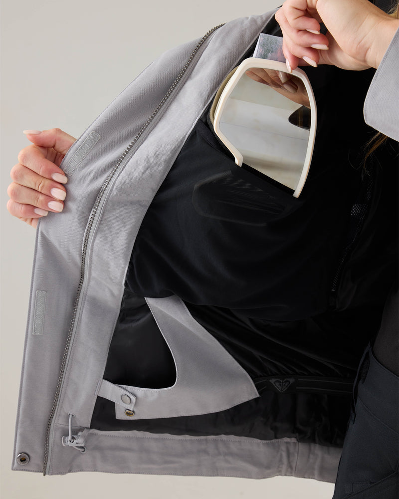 A person holds open the light gray Roxy Meade Jacket, revealing a black inner pocket where they are storing a pair of white ski goggles, highlighting the insulated warmth and waterproof protection this women’s snowboard jacket offers for your next adventure.