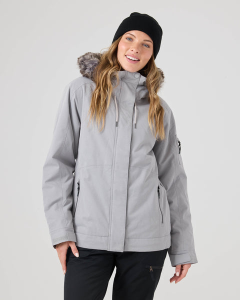 A woman smiles at the camera against a plain light background, wearing the Roxy Meade Jacket—a gray hooded winter coat with fur trim, insulated warmth, and waterproof protection—paired with black pants and a black beanie.