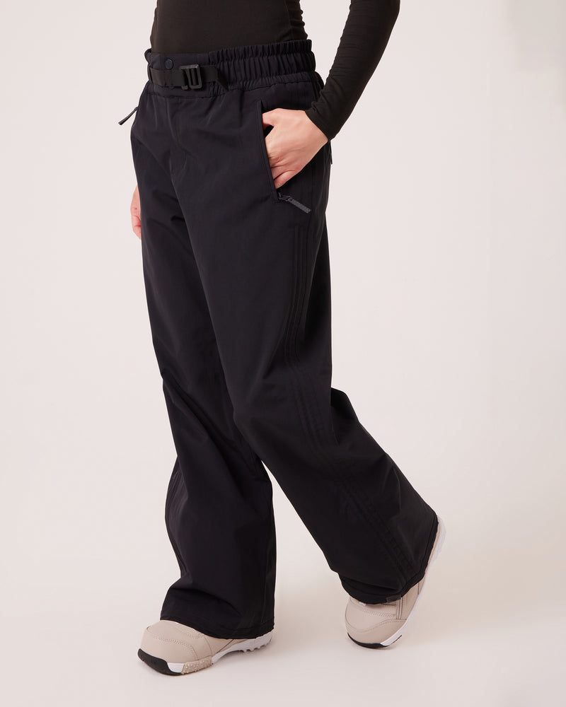 A person stands against a plain background with one hand in their pocket, wearing Roxy’s Bleeker Pants—wide-leg black pants made from PFAS-free fabric—paired with a black long-sleeve top and light-colored sneakers.