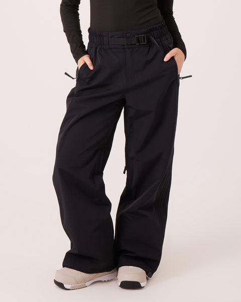 A person stands against a plain light background with hands in pockets, wearing the Bleeker Pants by Roxy—loose-fitting black pants inspired by women’s snowboard styles. The pants feature an elastic waistband, belt, and visible zippers for style and function. Crafted from waterproof-breathable materials with PFAS-free finishes, they provide both comfort and sustainability. The look is completed with a black long-sleeve top and white sneakers.