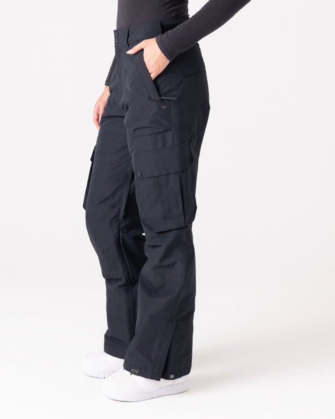 A person is wearing Roxy Trooper Cargo Pants in black, paired with a long-sleeve dark shirt and white shoes. The pants feature multiple pockets, and the individual stands with one hand tucked into a pocket against a plain, light-colored background.