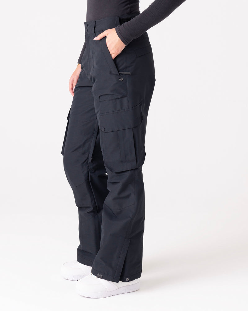 A person is wearing Roxy Trooper Cargo Pants in black, paired with a long-sleeve dark shirt and white shoes. The pants feature multiple pockets, and the individual stands with one hand tucked into a pocket against a plain, light-colored background.