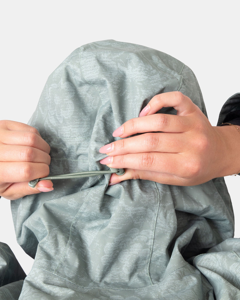 A person pulls the drawstring on the hood of a light gray Roxy Jetty 3N1 women’s snowboard jacket, tightening it with both hands so their face is mostly obscured by the hood.