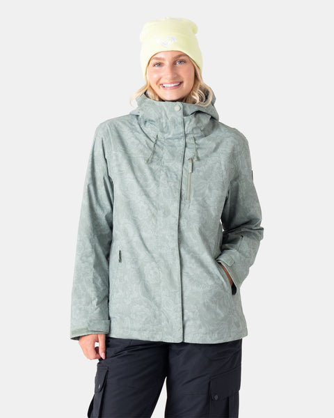 A woman stands smiling with one hand in her pocket, wearing the Roxy Jetty 3N1 Jacket in Olive Combo—an eco-friendly women’s snowboard jacket made from recycled materials—paired with black pants and a pale yellow beanie, against a plain light gray background.