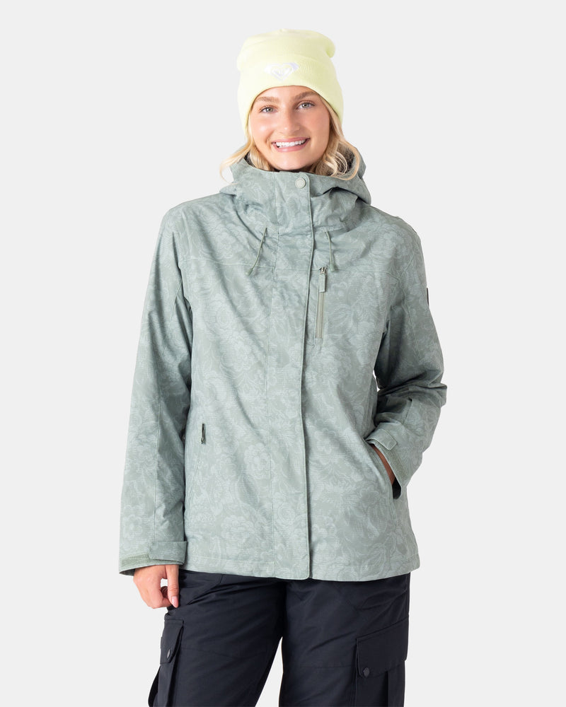 A woman stands smiling with one hand in her pocket, wearing the Roxy Jetty 3N1 Jacket in Olive Combo—an eco-friendly women’s snowboard jacket made from recycled materials—paired with black pants and a pale yellow beanie, against a plain light gray background.