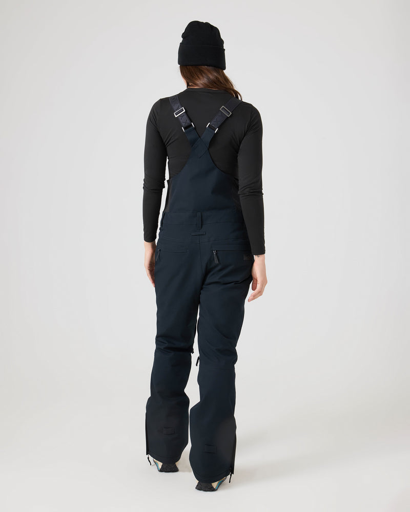 A person stands with their back to the camera, wearing a black beanie, long-sleeve shirt, and Roxy Rideout Bib overalls featuring crossed straps, set against a plain light background.