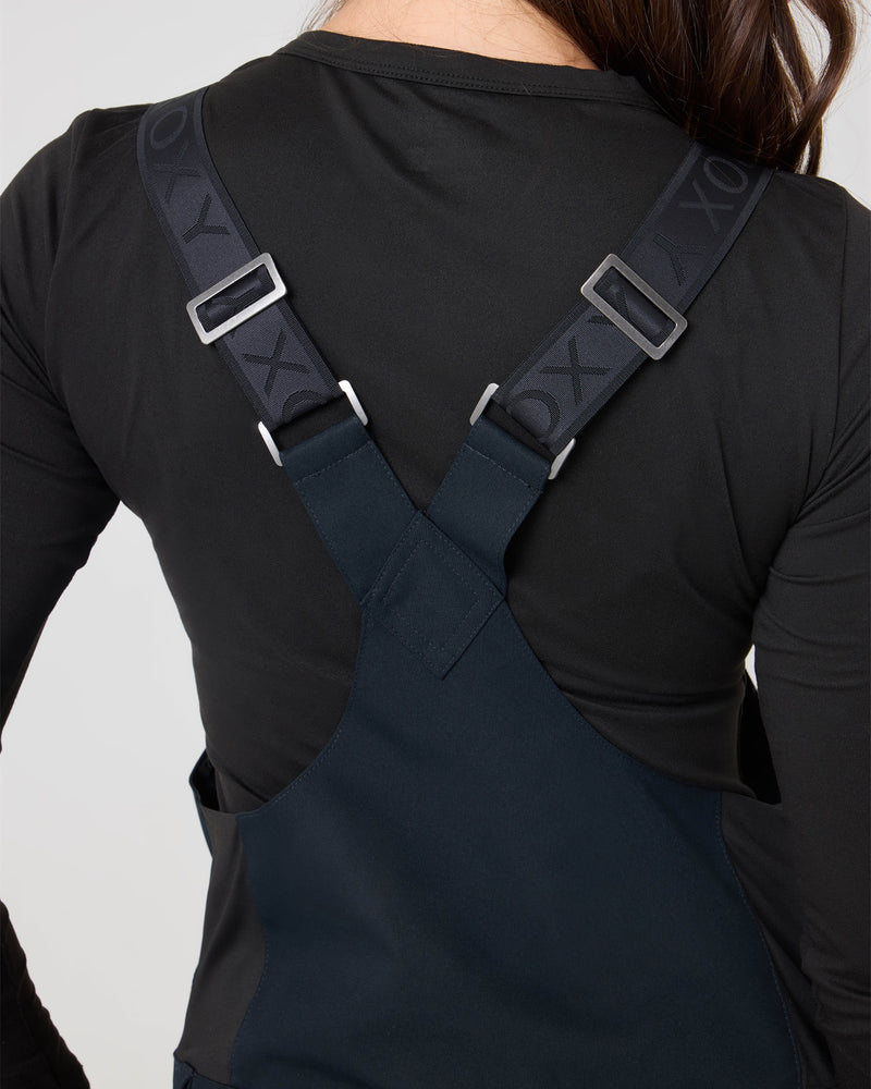 A person wearing a black long-sleeve shirt and navy Roxy Rideout Bib overalls, featuring lightly insulated material and adjustable straps marked with XOX. The image highlights the upper back and shoulders, showcasing the crossed straps of the Rideout Bib for a storm-ready look.