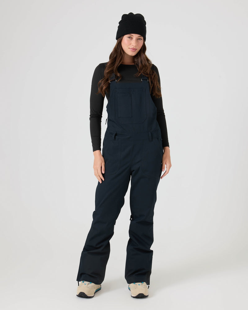 A person stands facing forward wearing a black beanie, a long-sleeve black shirt, and storm-ready black Roxy Rideout Bib overalls with light-colored sneakers against a plain white background.