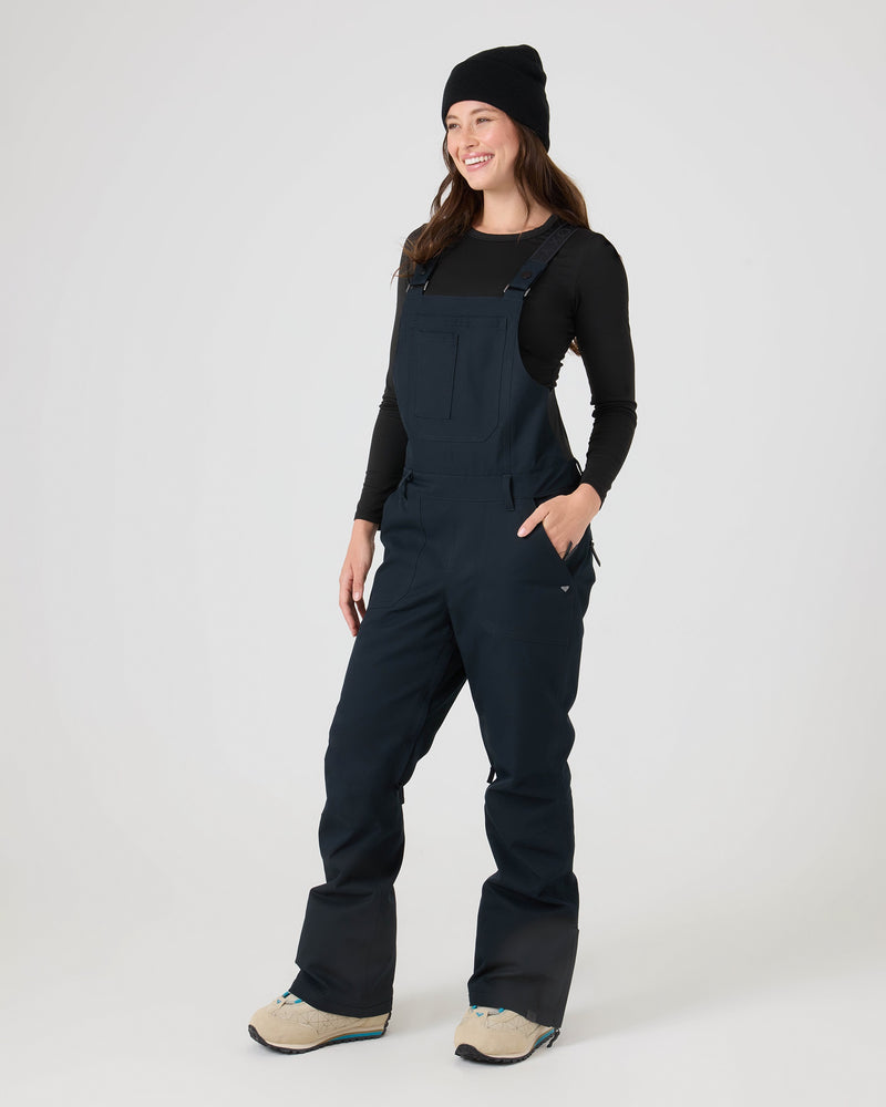 Clad in Roxy’s Rideout Bib—a storm-ready, lightly insulated overall—along with a black beanie and black long-sleeve shirt, a person stands smiling with their hands in their pockets against a plain light background.