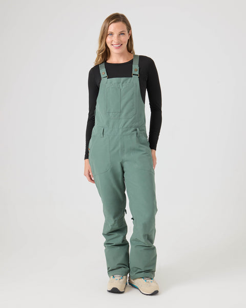 A woman stands smiling, wearing a black long-sleeve shirt and Roxy Rideout Bib overalls in Duck Green with large pockets and beige sneakers, against a plain white background.