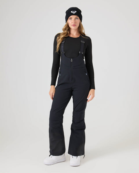 A woman wearing the Roxy Summit Bib overalls, paired with a black beanie, long-sleeve black shirt, and white boots, stands facing forward against a plain light background.