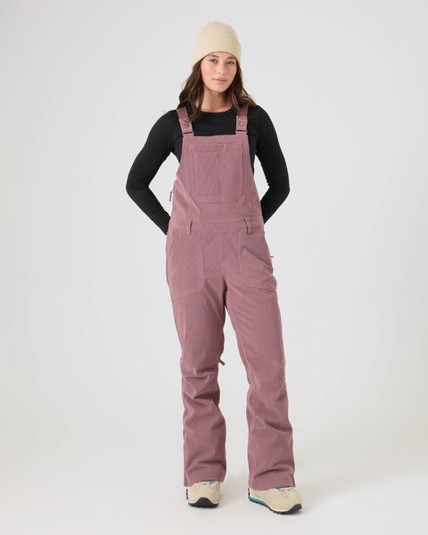 A woman wearing a beige beanie, a black long-sleeve shirt, and the Roxy Rideout Bib in Rose Taupe with PrimaLoft insulation stands with her hands in her pockets against a plain white background.
