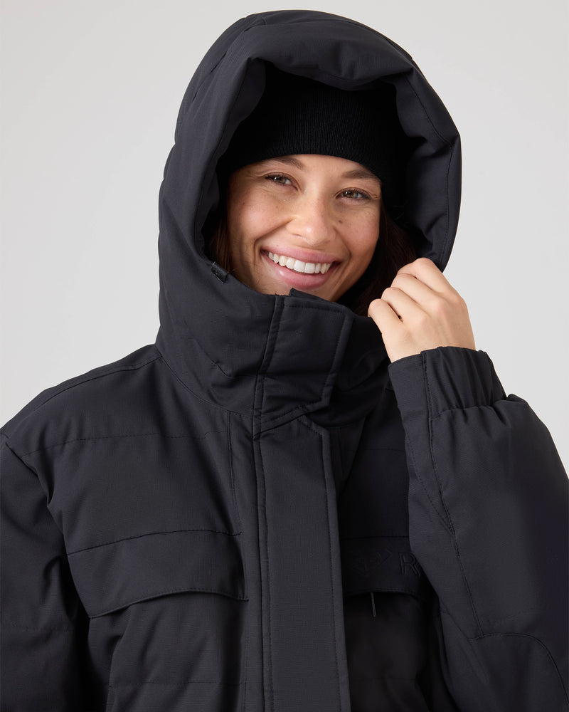A woman wearing the Roxy Jewelz Jacket—a black ski jacket with PrimaLoft insulation—pairs it with a black beanie, smiling broadly and holding the edge of her hood against a neutral, uncluttered background.