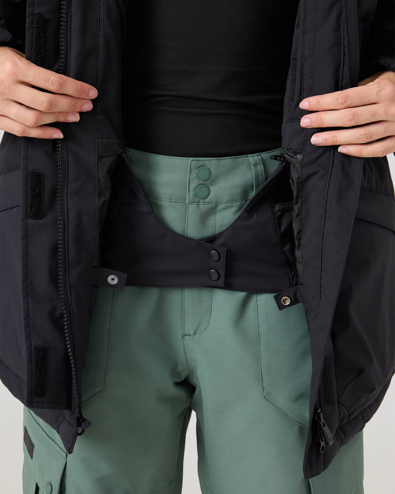 Person wearing green pants and a black jacket, holding the jacket open to reveal snap closures and the inner waistband details of the pants—ideal features for pairing with the Roxy Jewelz Jacket, which offers cozy warmth with PrimaLoft insulation.