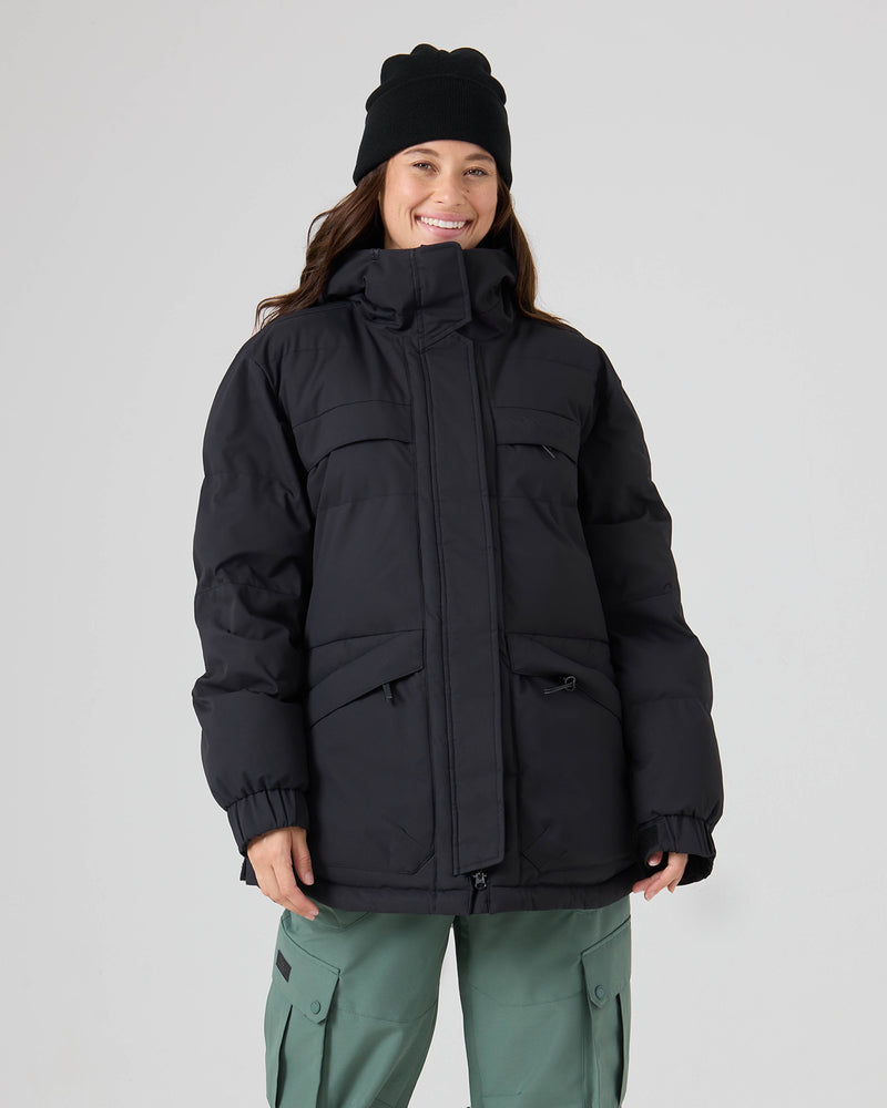 A person wearing the Roxy Jewelz Jacket in black with PrimaLoft insulation, green cargo pants, and a black beanie smiles while standing against a plain light gray background.