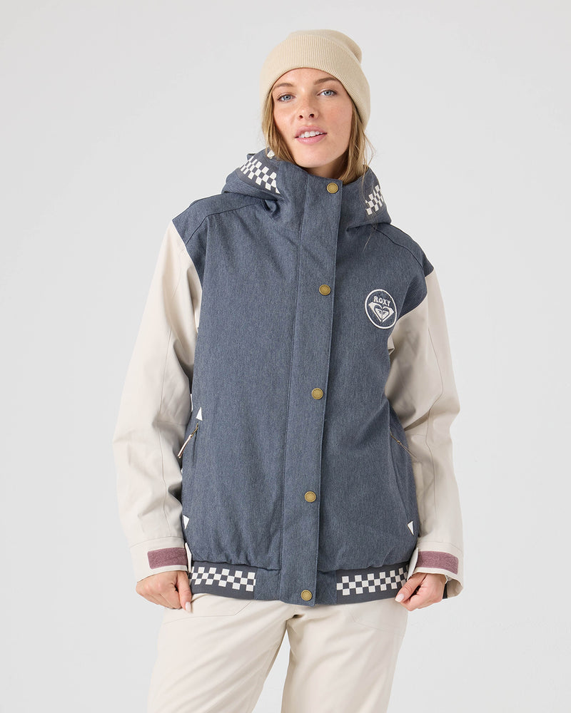A person wearing a beige beanie, beige pants, and the Roxy Cheer Up Jacket in Denim—which features blue and cream hues with checkered details—stands against a plain light background, gazing slightly to the side with a neutral expression. The technical outerwear from Roxy combines stylish accents with dependable protection from the elements.
