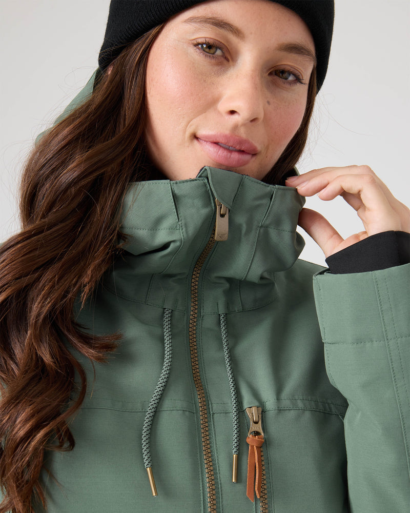 A person wearing the Roxy Free Love Jacket—a green zip-up women’s snow jacket with PrimaLoft insulation—and a black beanie looks at the camera, touching the collar of the jacket with one hand.