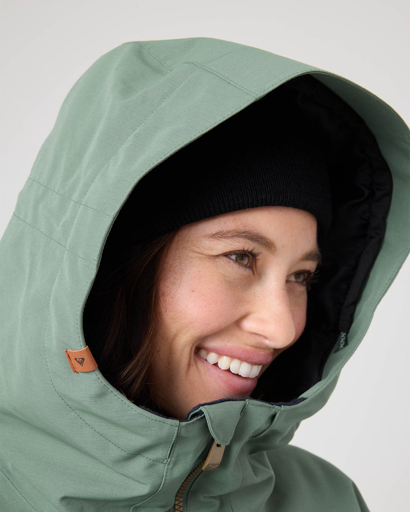 A person smiles while wearing the Roxy Free Love Jacket, a light green women’s snow jacket with PrimaLoft insulation, and a black beanie. The close-up image highlights their face and the textured fabric details of the jacket’s hood.