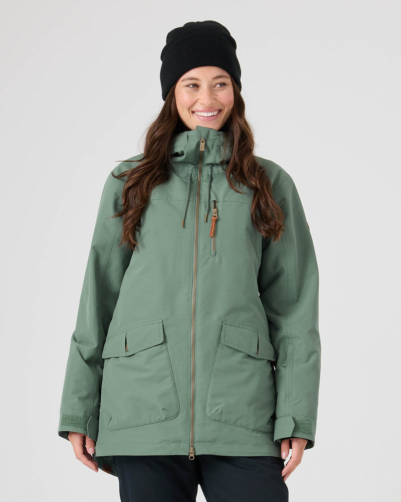A woman wearing the Roxy Free Love Jacket in Duck Green—with PrimaLoft insulation, a high collar, front zipper, chest pocket, two large flap pockets for 15K/10K performance—and a black beanie is smiling. She stands against a plain light gray background.