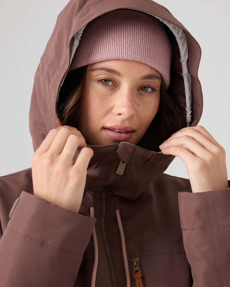 A person wearing the Roxy Free Love Jacket in mauve, featuring 15K/10K protection, and a matching knit beanie looks at the camera, holding the jacket hood with both hands.