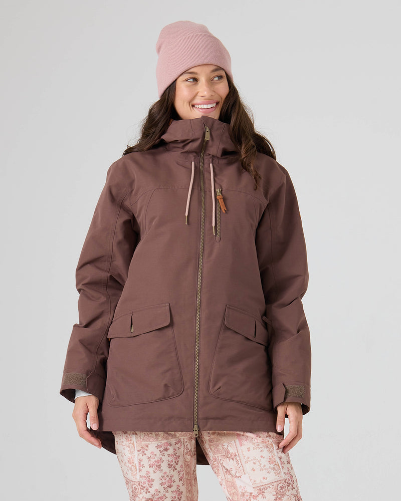 A woman smiles as she stands against a plain background, wearing the Roxy Free Love Jacket in the Shopping Bag color. Featuring PrimaLoft insulation for warmth and dependable 15K/10K protection from the elements, she pairs her jacket with light pink floral-patterned pants and a pink knit beanie, her hands relaxed by her sides.
