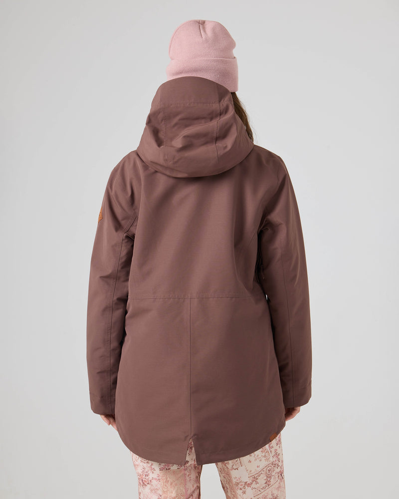 A person is seen from behind against a plain light background, wearing the Roxy Free Love Jacket — a mauve women’s snow jacket with PrimaLoft insulation — styled with a pink knit beanie and light pink patterned pants.