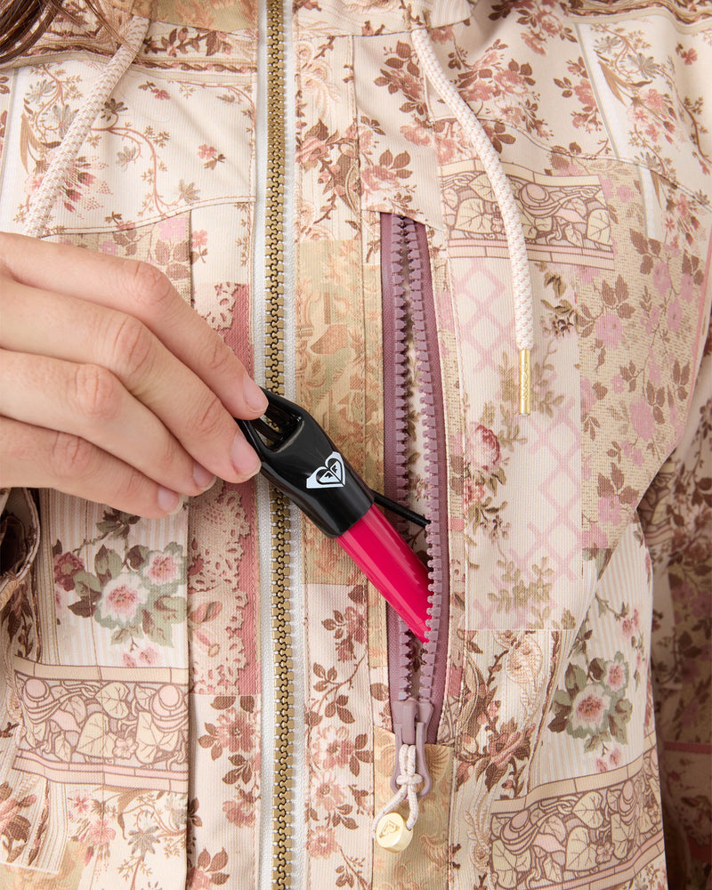 A person holds a black and red lipstick near the zipper pocket of a floral-patterned women’s snow jacket, appearing to place it inside—highlighting both the stylish design and practical features of the Roxy Incline Jacket.