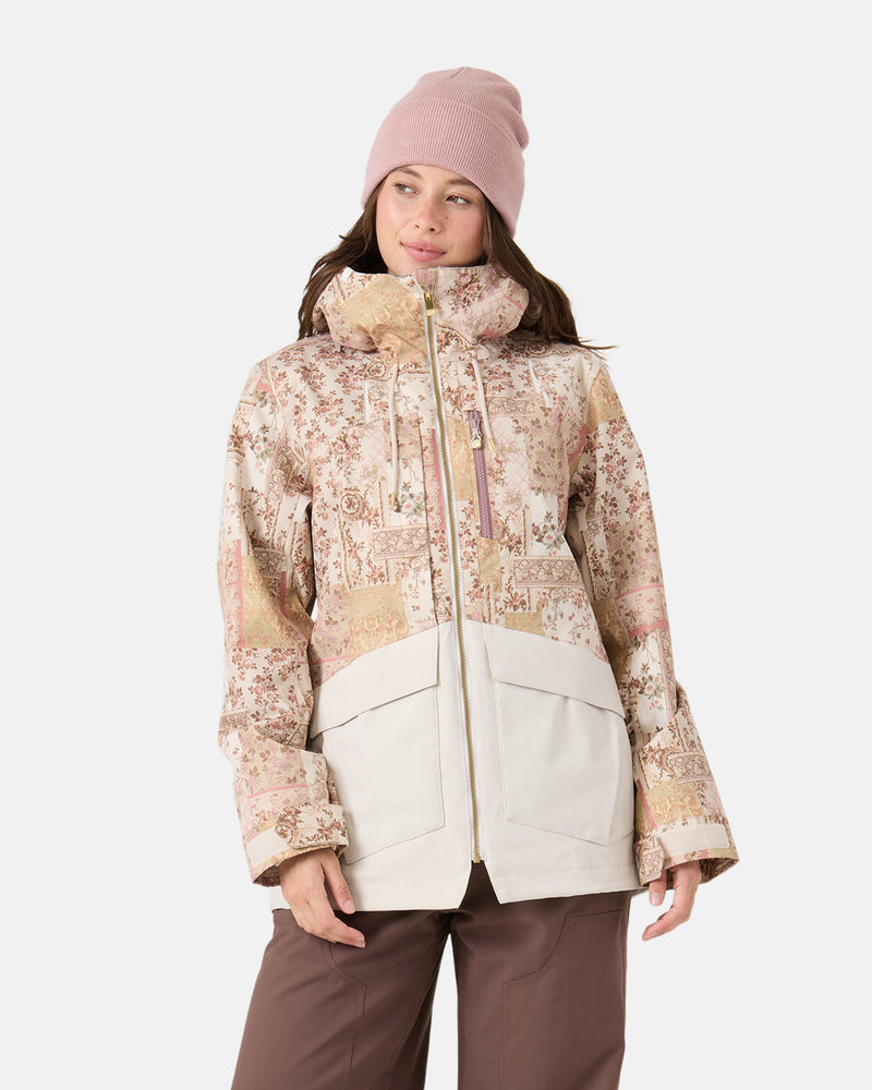A woman wearing a light pink beanie, brown pants, and the Roxy Incline Jacket—a beige and pink patterned women’s snow jacket featuring large pockets and 15K protection—stands against a plain white background.