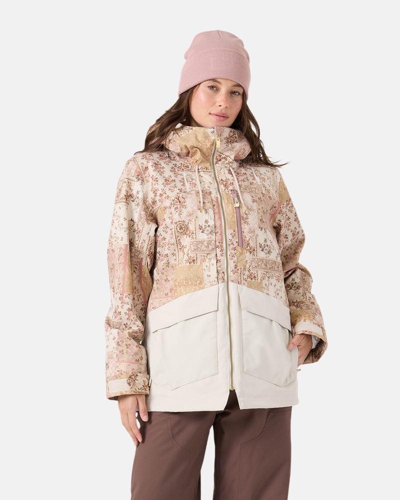 A woman in a pink beanie and brown pants stands against a white background wearing the Roxy Incline Jacket in Earthy Wahine, featuring a floral pattern, large cream pockets, and 15K protection.