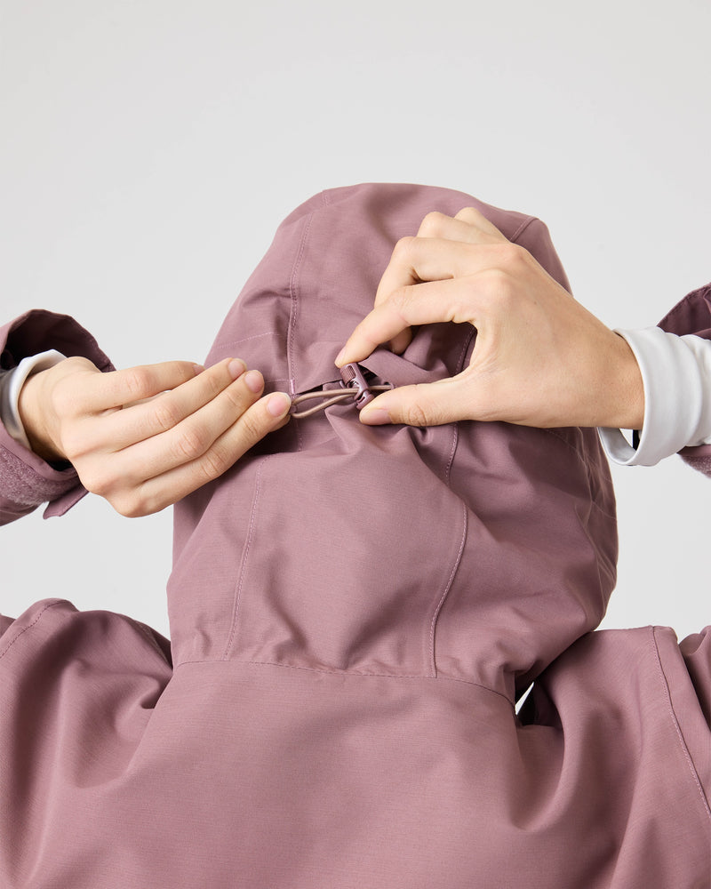 A person adjusts the drawstring on the back of a mauve Roxy Incline Jacket, shown from behind. Their hands pull on the toggle to tighten or loosen the hood of this women’s winter jacket, which features 15K protection. The background is plain and light-colored.