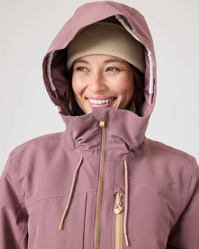 A person is smiling and looking slightly to the side while wearing the Roxy Incline Jacket, a mauve women’s snow jacket with 15K protection, zip closure, and drawstrings, paired with a beige beanie. The background is plain and light-colored.