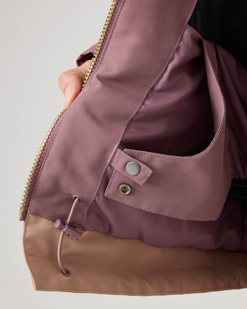 A close-up of a hand holding open the Roxy Incline Jacket in mauve and tan, highlighting the interior pocket with a snap button and an adjustable drawstring at the hem—thoughtful touches that keep you organized and cozy in this women’s snow jacket featuring 15K protection.