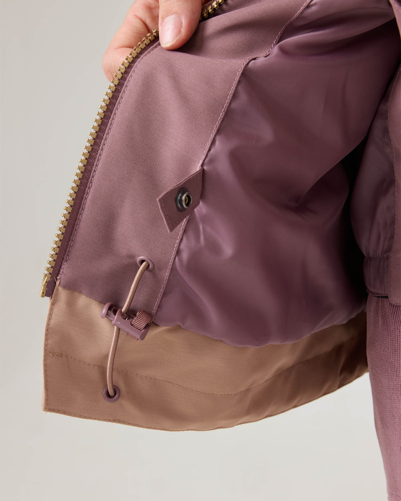 A hand holds open the inside of the mauve-colored Roxy Incline Jacket, revealing a drawstring, grommets, and a button on the interior lining near the zipper—features designed to provide warmth and 15K protection for winter adventures.