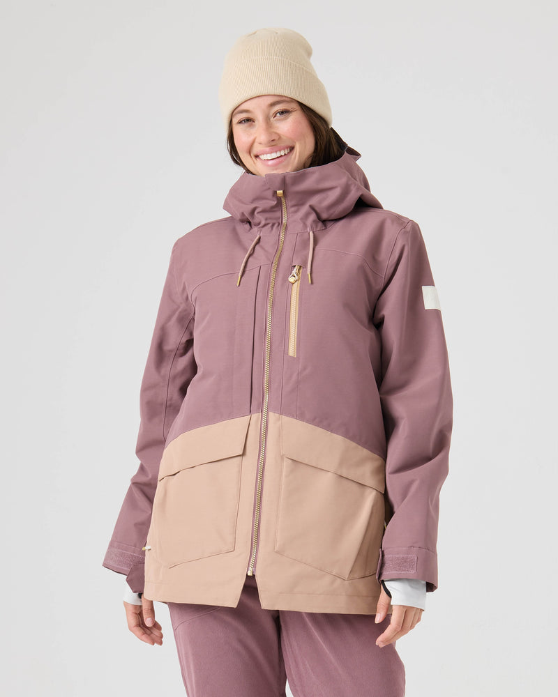 A person wearing the Roxy Incline Jacket in Rose Taupe, paired with matching mauve pants and a beige beanie, smiles while standing against a plain light background.
