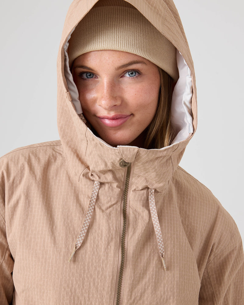 A person wearing the Roxy Downtown Anorak—a beige, recycled nylon hooded jacket with drawstrings—alongside a beanie, looks into the camera with a slight smile. The jacket is zipped up, and the background is plain and light-colored.