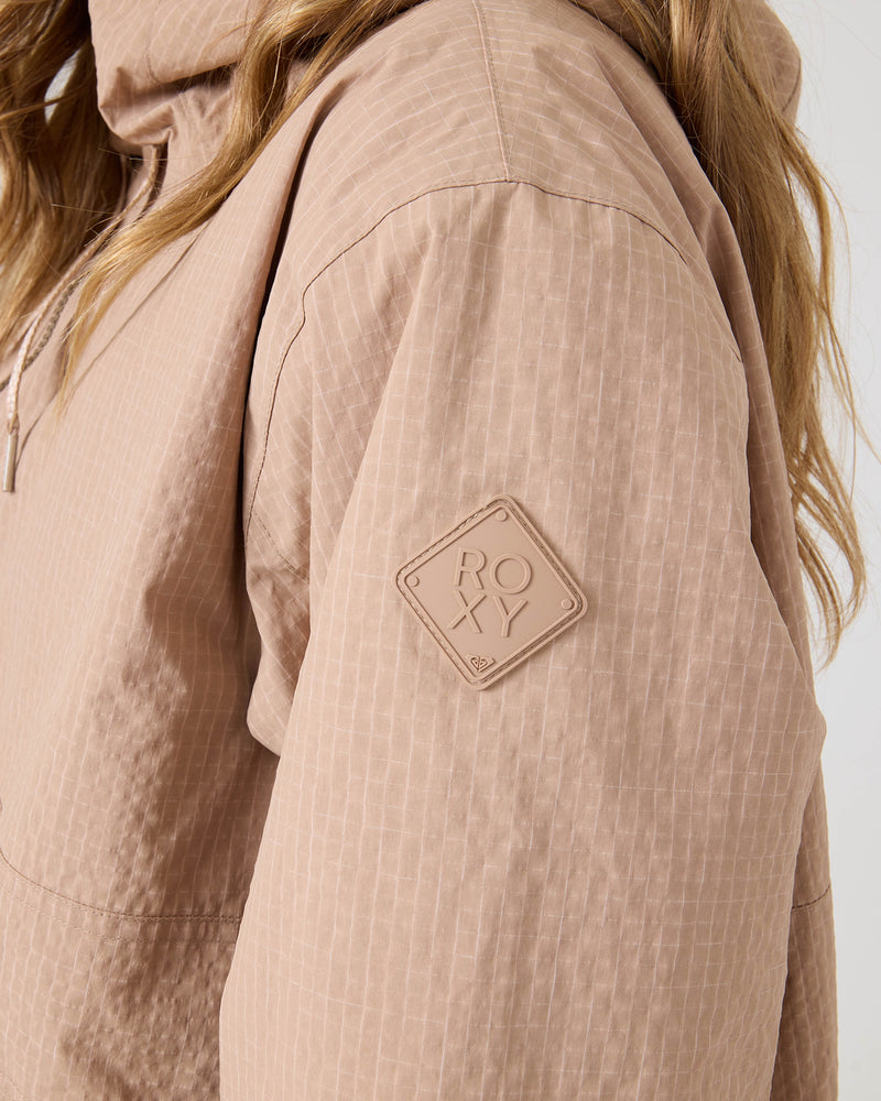A close-up of a person wearing the ROXY Downtown Anorak, a light brown textured women’s snowboard jacket featuring a diamond-shaped ROXY patch on the sleeve. Only the upper body and part of the hair are visible, showcasing the quality recycled nylon material.