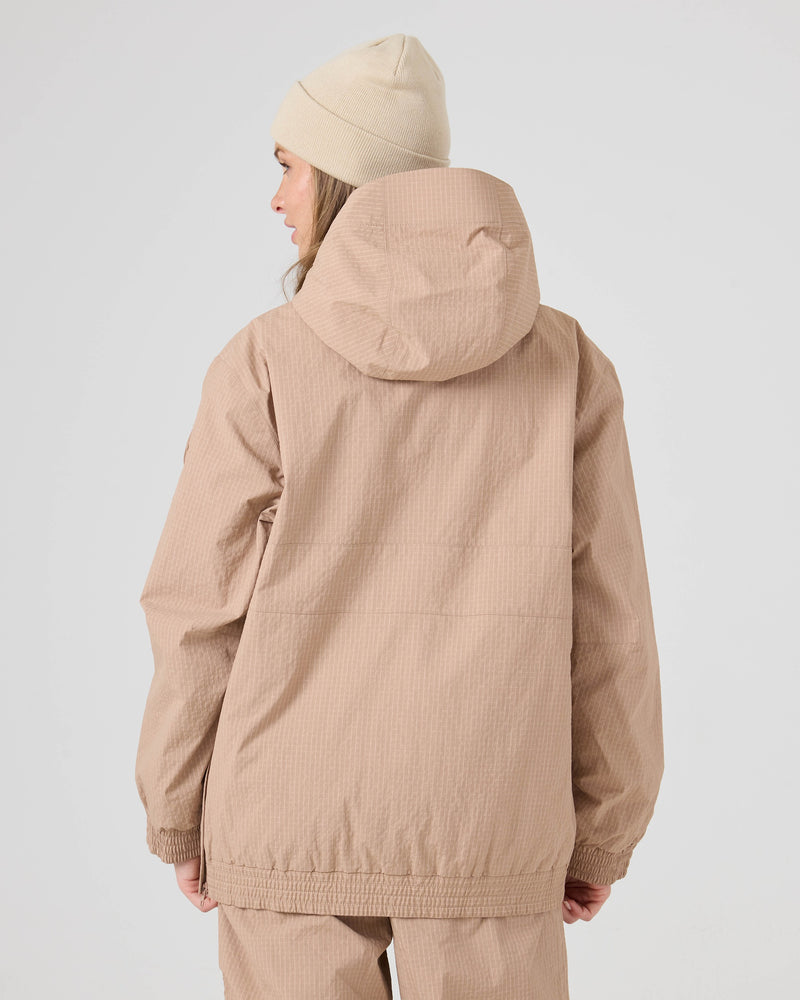 A person wearing the Roxy Downtown Anorak, a beige women’s snowboard jacket made from recycled nylon, along with matching pants, stands facing away from the camera. They have long hair and are also wearing a cream-colored beanie. The Downtown Anorak stands out against the plain, light-colored background.