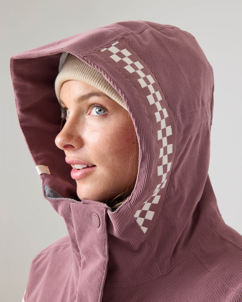 A woman in the Roxy Cheer Up Jacket—a mauve women’s snowboard jacket with a checkered-pattern hood made from recycled polyester—looks to the side, wearing a beige beanie underneath and smiling softly.