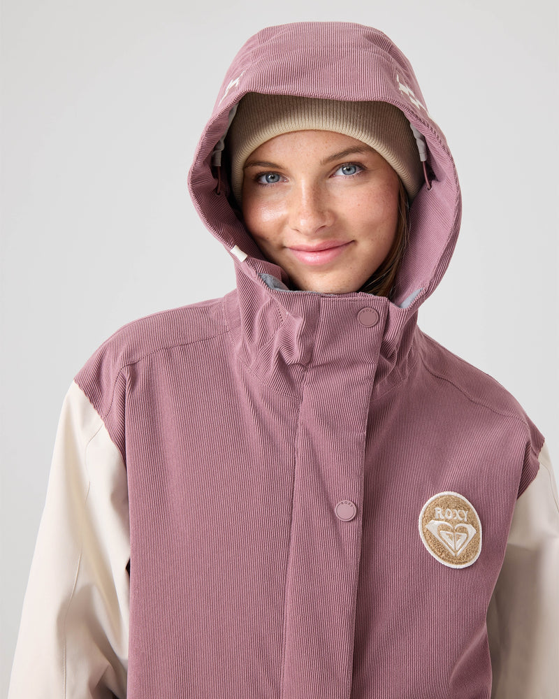 A person wearing the Roxy Cheer Up Jacket—a mauve women’s snowboard jacket with a varsity style and ROXY patch, crafted from recycled polyester—pairs it with a beige beanie and smiles softly at the camera against a plain background.