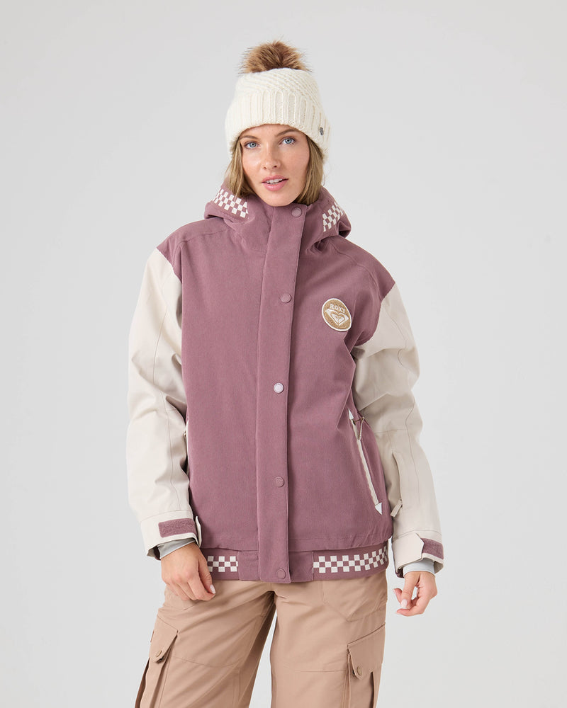 A woman wearing the Roxy Cheer Up Jacket in Rose Taupe—a mauve and beige varsity-style piece made from recycled polyester—paired with tan cargo pants and a white knit beanie with a brown pom-pom, standing against a plain light background.