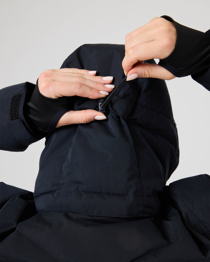 A person zips up the hood of a dark Roxy Snowdrift Jacket, revealing only their hands and upper back. Crafted from recycled polyester and insulated with faux down, the Roxy Snowdrift Jacket features thumb holes in the sleeves. The background is plain and light-colored.