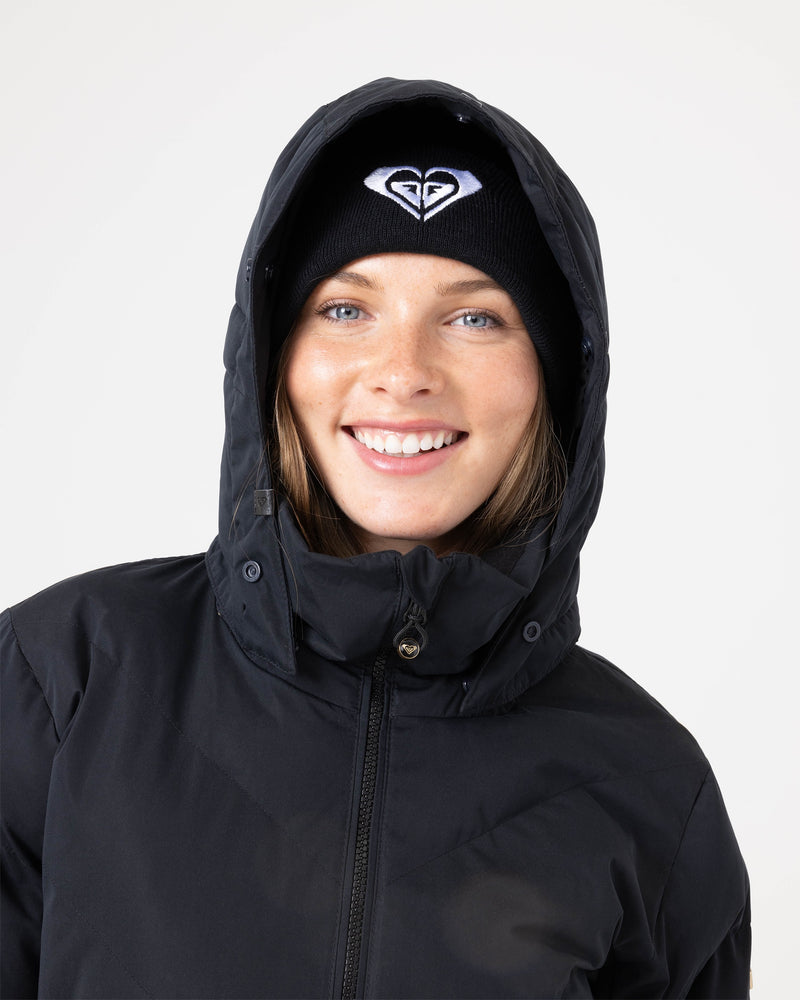A woman smiles while wearing the Roxy Snowdrift Jacket in black, crafted from recycled polyester and insulated with faux down. She pairs it with a black beanie adorned with a white heart-shaped logo on the front. Her light-colored eyes and brown hair peek out from beneath the beanie.