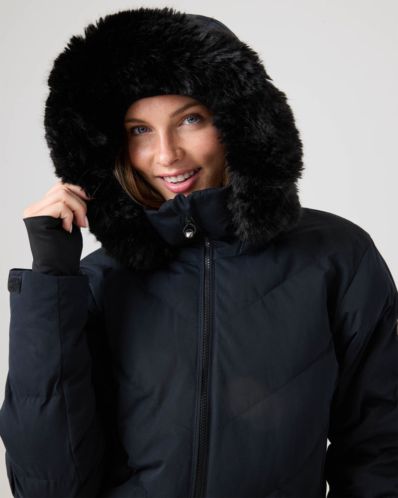 A woman smiles while wearing the Roxy Snowdrift Jacket, a black coat with a fluffy fur-lined hood made from recycled polyester faux down, as she holds the edge of the hood against a plain light background.