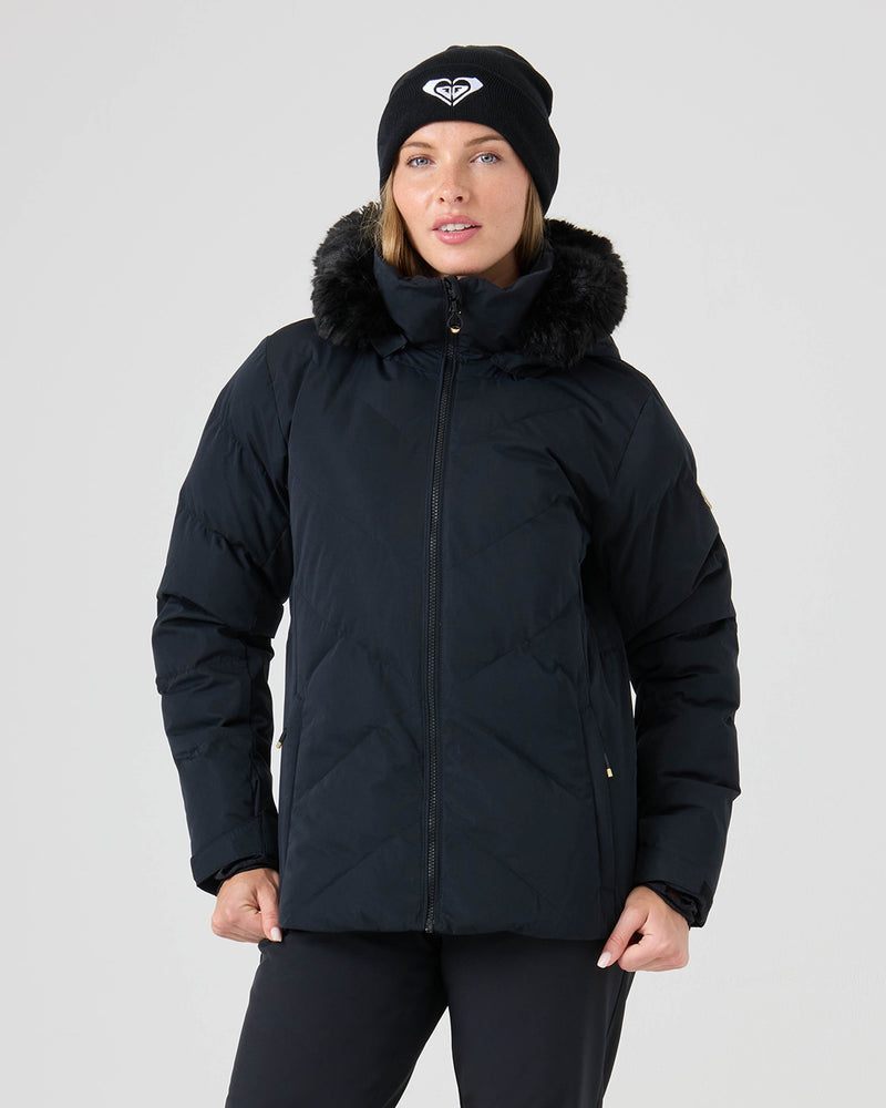 A woman stands against a plain light background wearing the Roxy Snowdrift Jacket in black, featuring a fur-lined hood made from faux down and recycled polyester, paired with a matching black beanie.