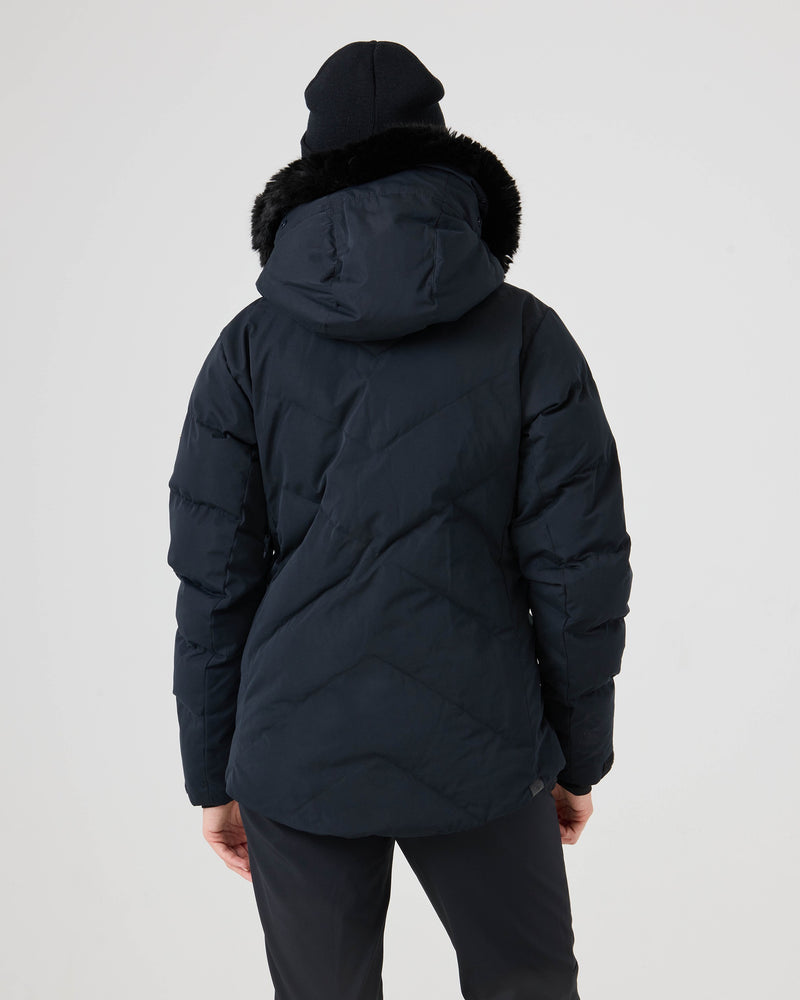 A person stands facing away, dressed in the Roxy Snowdrift Jacket—a dark winter coat with a hood trimmed in black faux fur, made from recycled polyester—paired with a dark beanie and dark pants, set against a plain light background.