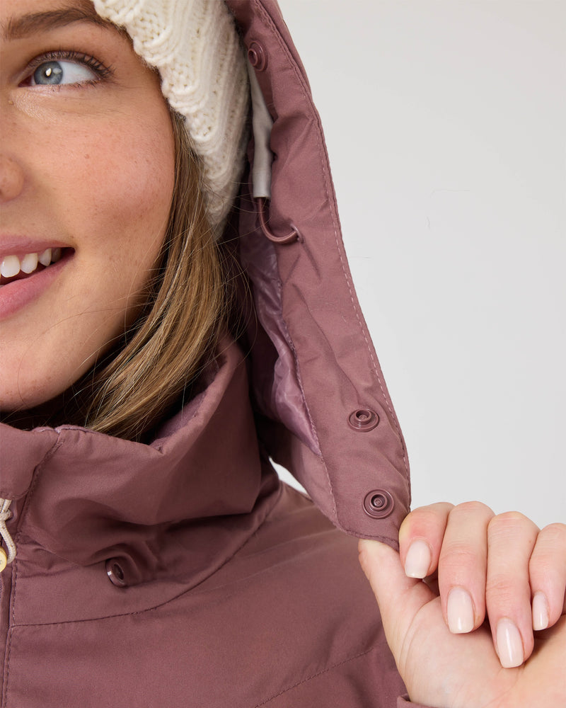 A woman wearing the Roxy Snowdrift Jacket, crafted from recycled polyester and insulated with warm faux down, smiles as she holds the hood with one hand beneath a white knit beanie, revealing part of her face.