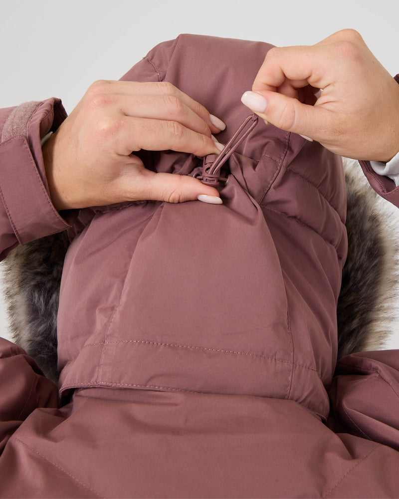 A person tightens the hood drawstring on a mauve Roxy Snowdrift Jacket using both hands. Made from recycled polyester and insulated with soft faux down, this outerwear by Roxy combines sustainability with style.