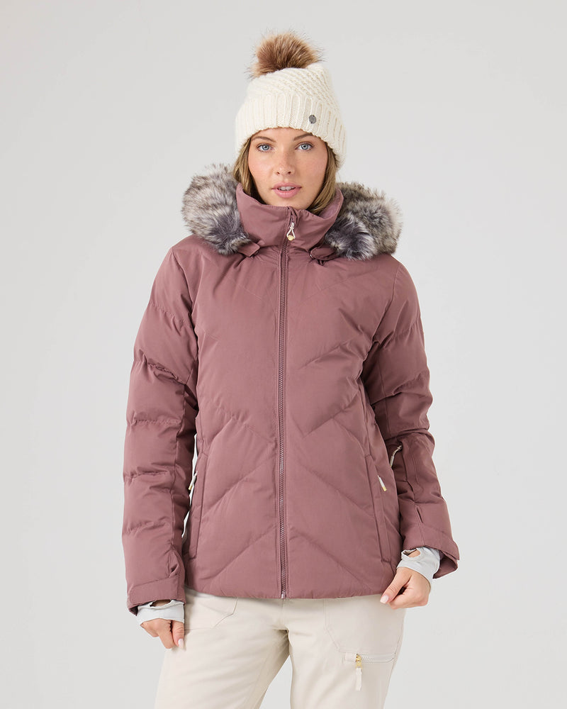 A woman wearing a rose taupe Roxy Snowdrift Jacket made from recycled polyester with faux down insulation, featuring a fur-lined hood and a white knit beanie with a pom-pom, stands against a plain light background, facing forward with a neutral expression.