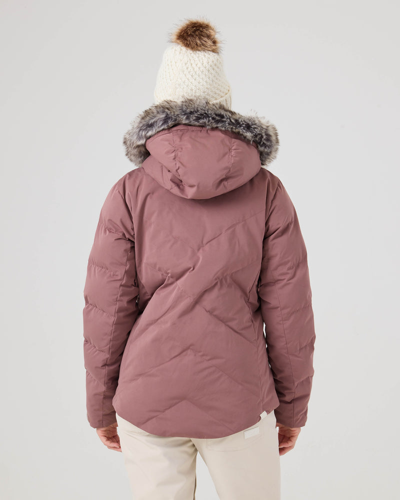 A person wearing the Roxy Snowdrift Jacket in mauve, featuring a recycled polyester shell, faux down insulation, and a fur-lined hood, paired with a white knit hat with a brown pom-pom and white pants, stands with their back to the camera against a plain background.