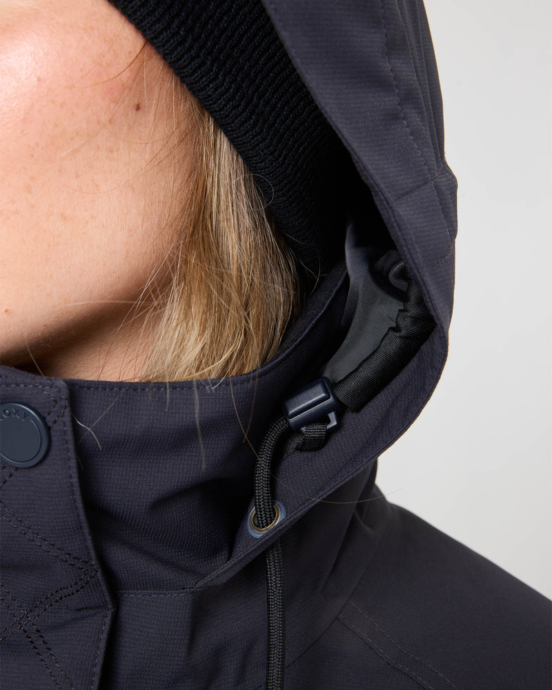 Close-up of a person wearing the Roxy Billie Jacket, featuring visible drawstring and button details, along with a black knit beanie. Part of the person’s blonde hair and cheek can also be seen.
